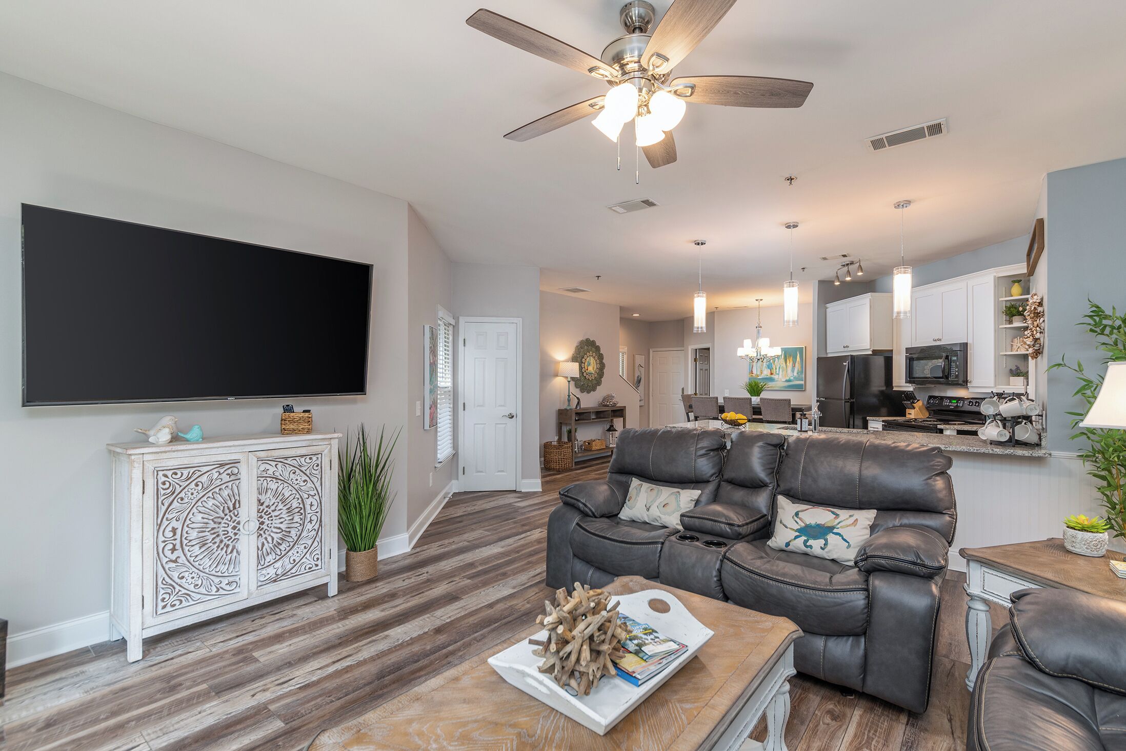 A spacious living room with a flat-screen TV, a leather sofa, a wooden coffee table, and a ceiling fan.