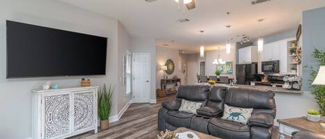 A spacious living room with a flat-screen TV, a leather sofa, a wooden coffee table, and a ceiling fan.