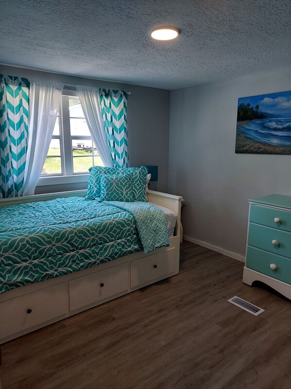 A bedroom with a bed, a dresser, a window with curtains, and a painting on the wall. - Small Town Florida (Brooksville)