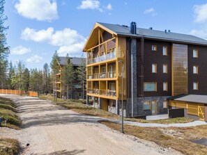 A multi-story building with balconies, a dirt road in front, and a forested area.