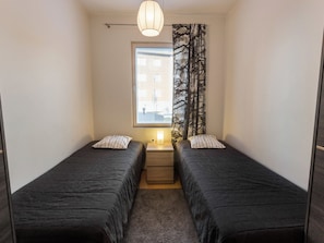 Ein kleiner, kompakter Raum mit zwei Einzelbetten, einem Nachttisch mit Lampe, einem Fenster mit Jalousien und einem Vorhang mit Baumotiv.