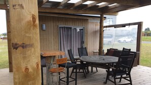 An outdoor wooden deck with a round table and chairs, a bar area, and a view of a parking lot. - Perfect in Pataua (Pataua North)