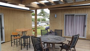 A covered outdoor patio with a wooden table and chairs, a sliding glass door, and a view of a backyard with a barbecue grill. - Perfect in Pataua (Pataua North)