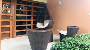 Una silla colgante con cojín, una silla de mimbre y una planta en maceta en un espacio exterior con puerta corrediza de vidrio y una pared con interruptor de luz.