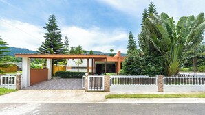 Una casa moderna con porche cubierto, entrada protegida por un portón y exuberante vegetación al fondo.