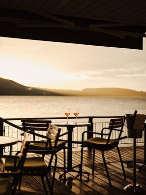 Une terrasse en bois avec une table et des chaises, donnant sur une étendue d’eau au coucher du soleil.