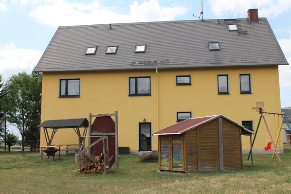 A yellow building with a grey roof, multiple windows, a small wooden shed, and a swing set. - Einzelzimmer Nr 4 - Pension Monika (Pausa/Vogtland)