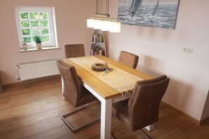 Un coin repas avec une table en bois, des chaises marron, un luminaire central, un radiateur, une fenêtre avec une plante et une bibliothèque.