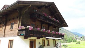 Una cabaña de madera tradicional con balcón decorado con flores, varios ventanales y un paisaje montañoso de fondo.