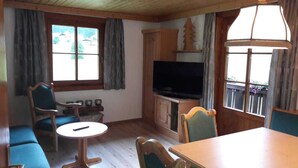 Een kamer met een televisie, houten meubels, een tafeltje en grote ramen.
