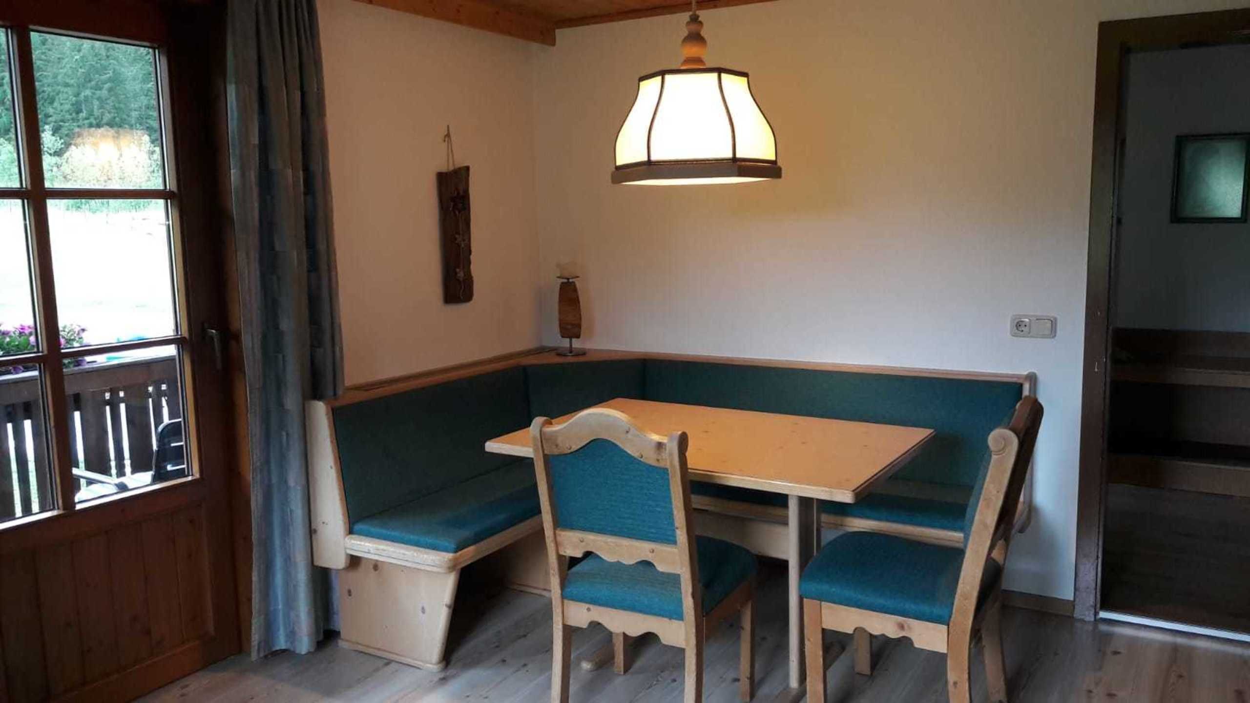 Una habitación con una mesa y sillas de madera, un banco, una lámpara colgante y una ventana que da a vistas de follaje.