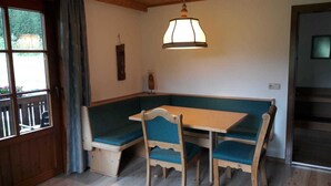 Een kamer met een houten tafel en stoelen, een bank, een hanglamp en een raam met uitzicht op groen.