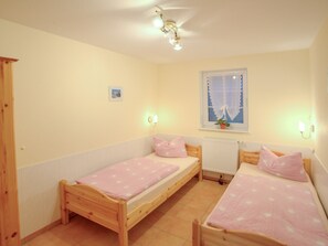 A bedroom with two wooden beds, each with a pink bedspread and matching pillows. There is a window with a lace curtain and a small potted plant on the windowsill. The room has white walls and a ceiling light fixture. - Vacation apartment 3 - Villa Lisa (Glowe)