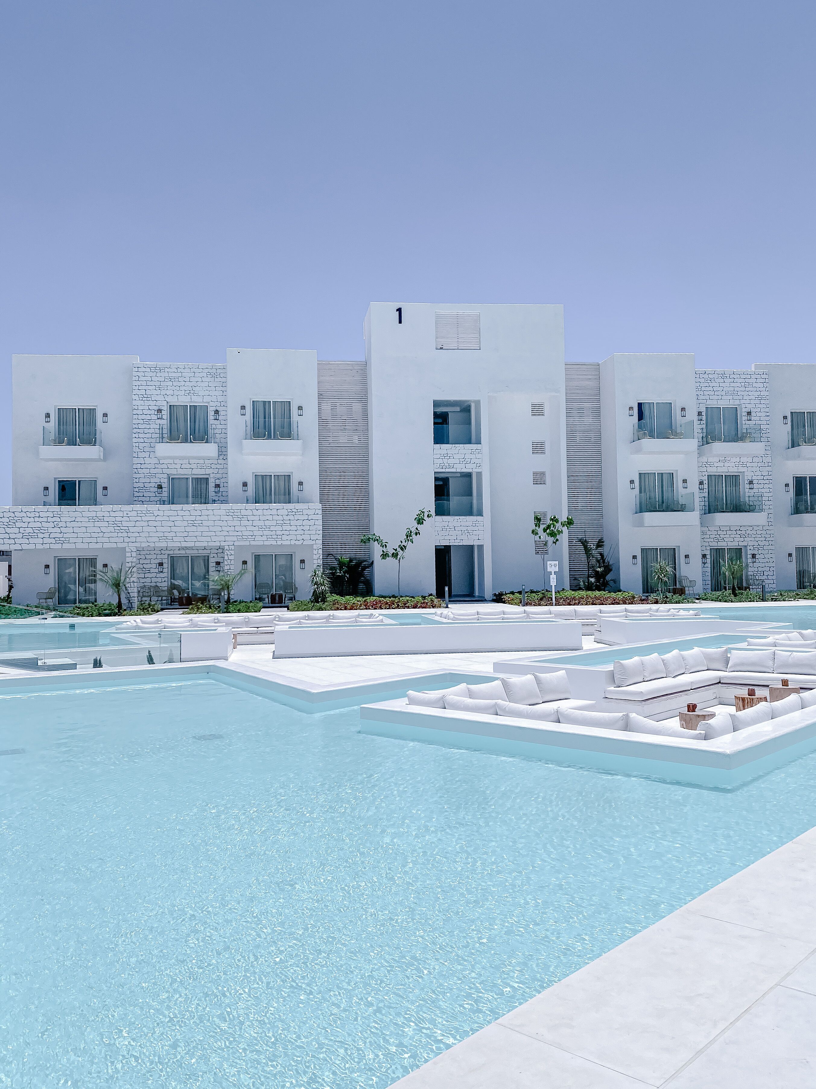 Un hotel moderno con zona de piscina, con edificios blancos con balcones y un cielo azul despejado.