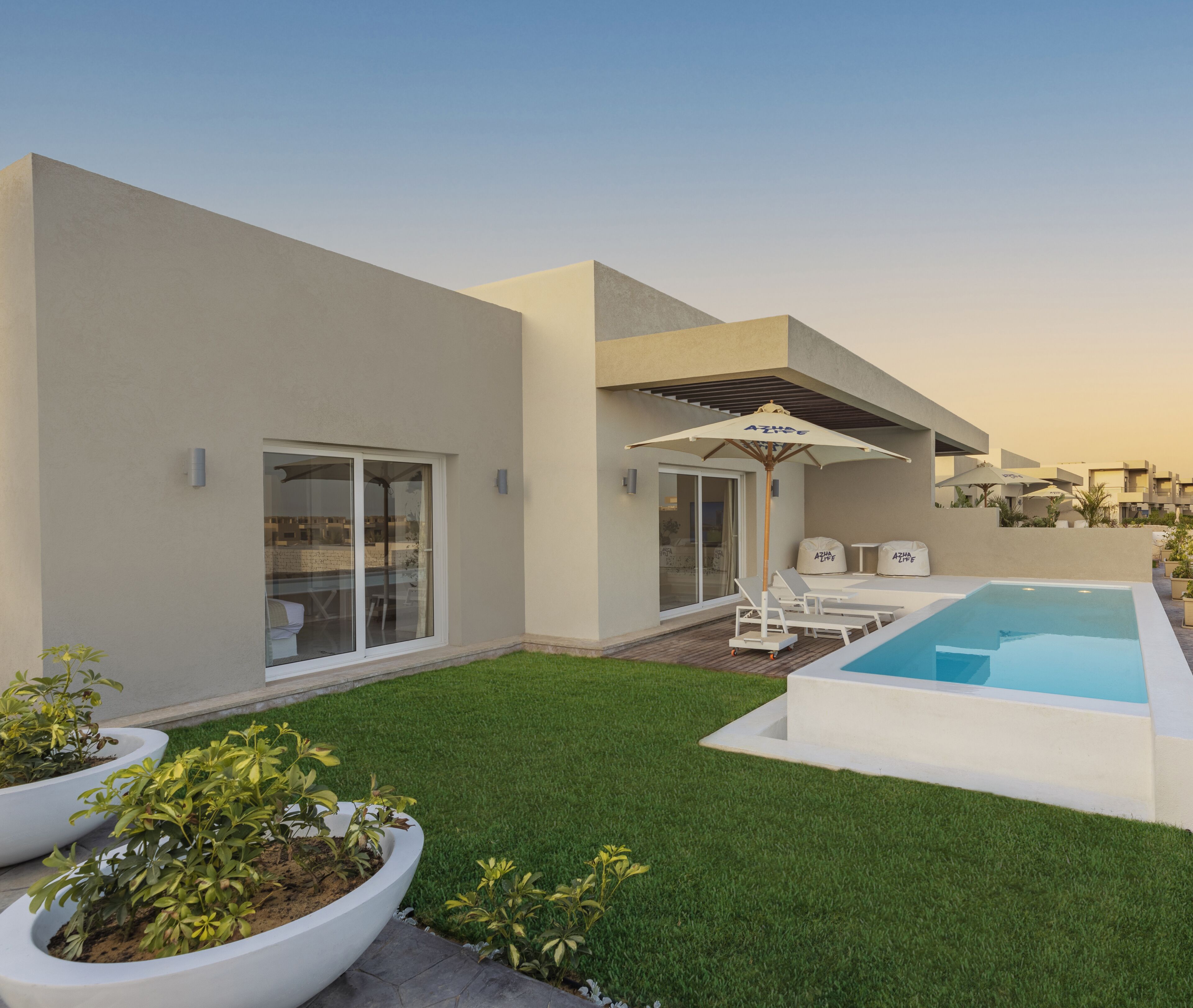 A modern outdoor pool area with a clear sky, a well-maintained lawn, and a seating area with lounge chairs and umbrellas.