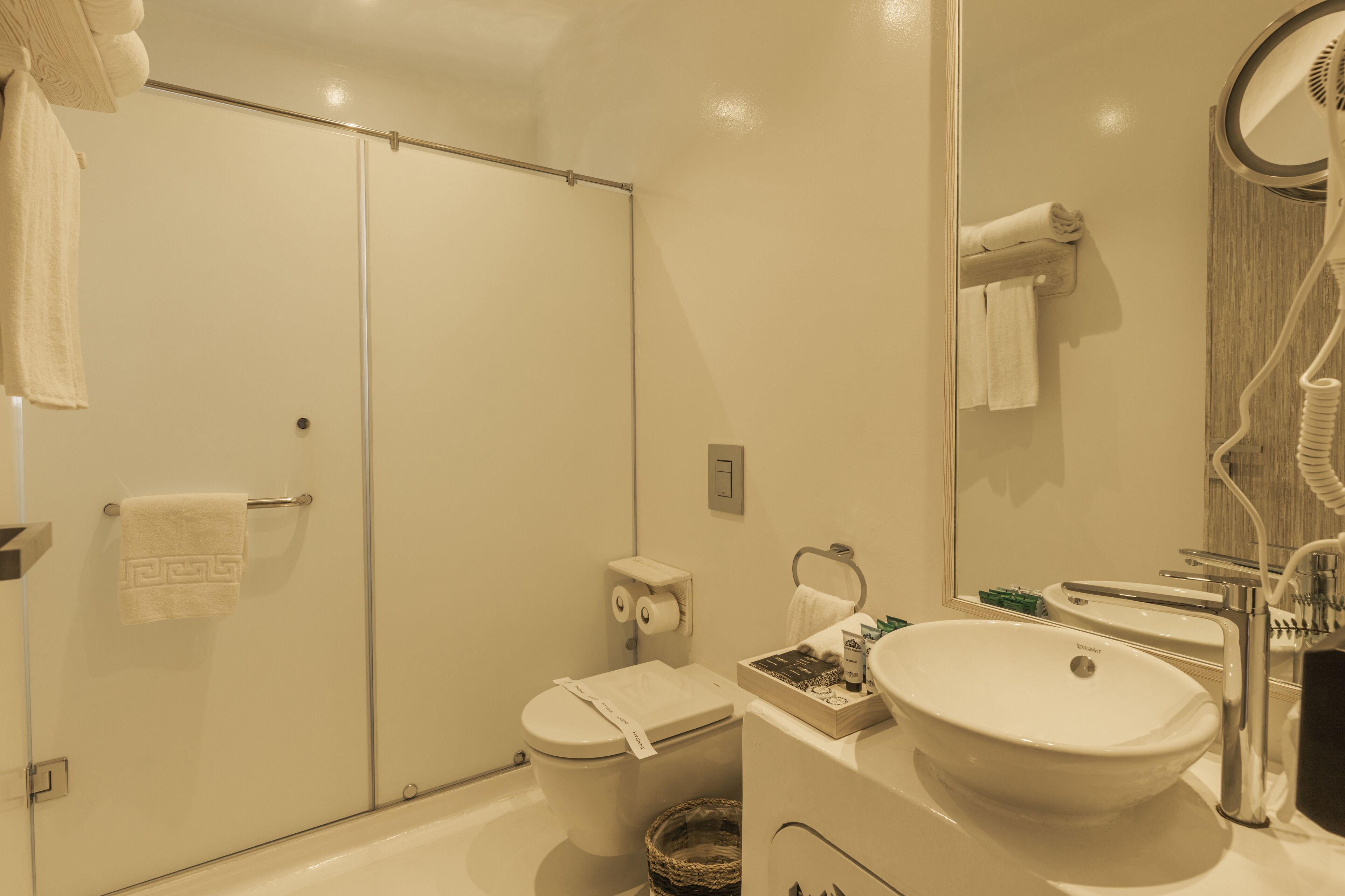 Un baño con cabina de ducha de vidrio, inodoro, lavamanos con espejo y secador de pelo.