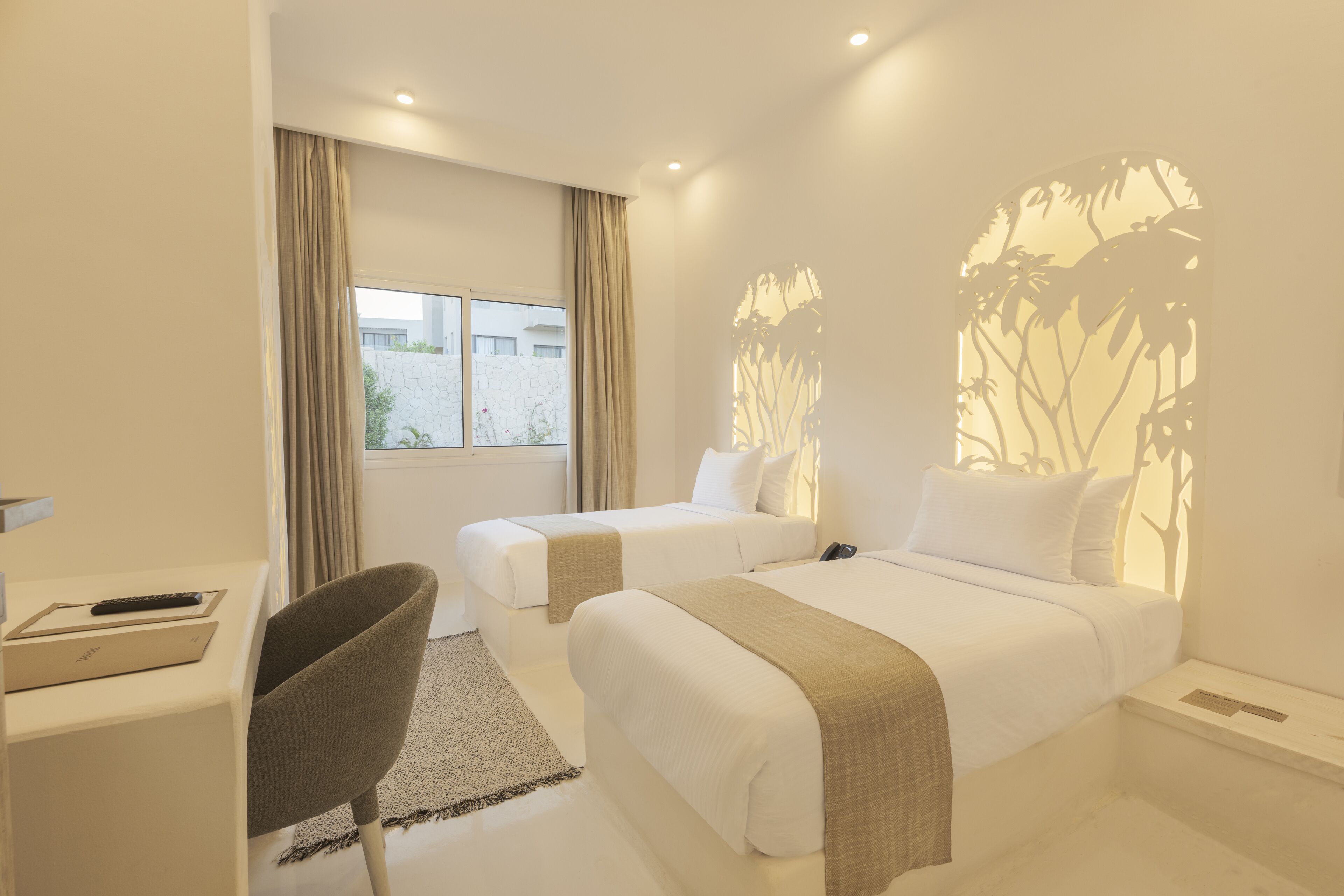 A hotel room with two beds, a desk with a remote control, a chair, and a view of the outside.