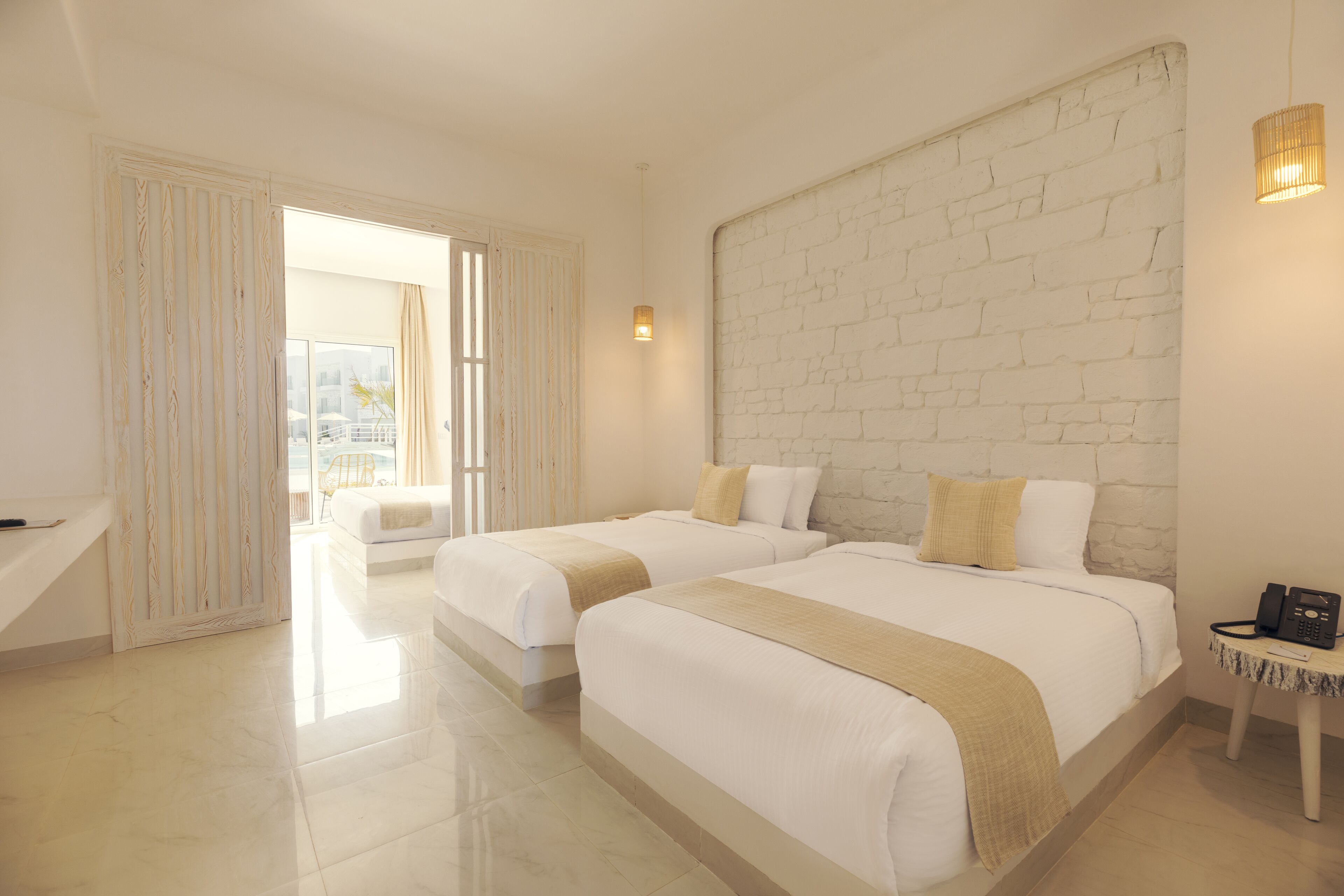 A hotel room with two beds, a white brick wall, a small table with a phone, and a view of the outside.