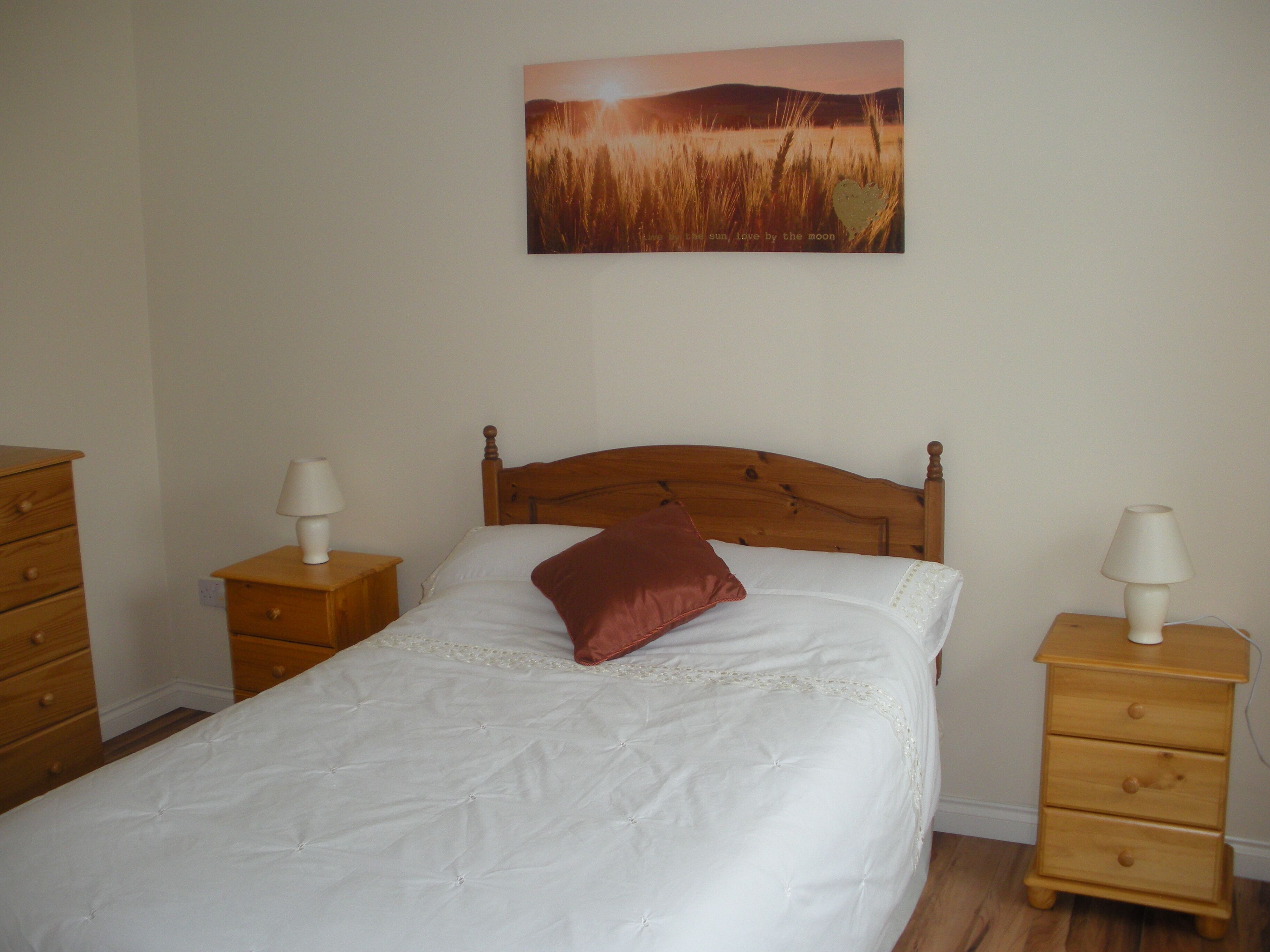 Un dormitorio con una cama de madera, ropa de cama blanca, una almohada roja, dos mesitas de noche con lámparas y un cuadro en la pared que muestra un atardecer sobre un campo.