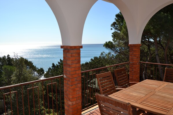 Un balcón con mesa y sillas de madera, con vistas a la costa, árboles y un cielo despejado.