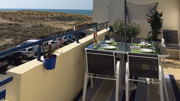 A balcony with a table and chairs, a blue umbrella, and a view of the beach and ocean. - Appart: T3 Front DE MER -orienté SUD - Terrasse 22 M2-2 Piscines- Parking (lacanau océan)