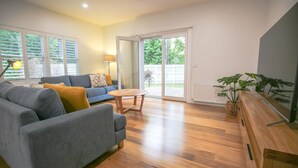 A spacious living room with a grey sofa, a wooden coffee table, and a large sliding glass door leading to a backyard. - BRIGHTER STAYS: Bright (Bright)