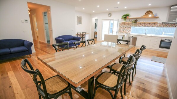 A spacious dining area with a large wooden table and six chairs, a blue sofa, and a kitchen in the background. - BRIGHTER STAYS: Bright (Bright)