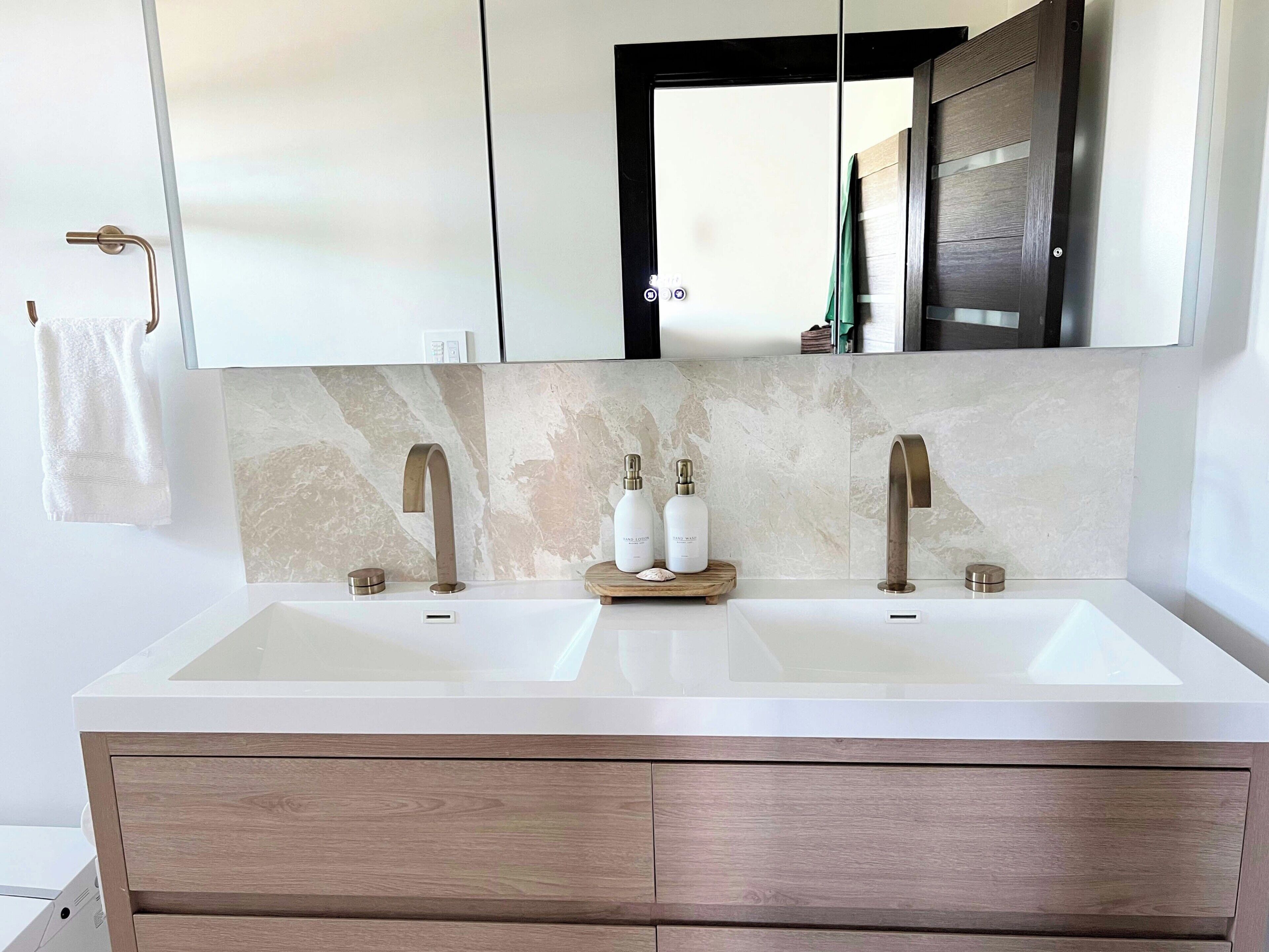Een moderne badkamer met een groot wit wastafel, twee gouden kraan en een houten wastafelmeubel met lades.