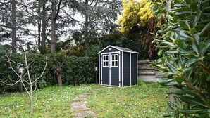 Um pequeno abrigo num jardim, com um caminho de pedra a levá-lo até ele, rodeado por vegetação e árvores.