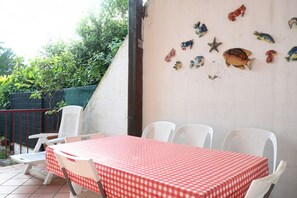 Een terras met een tafel, bedekt met een rood-wit geruit tafelkleed, omringd door witte plastic stoelen.