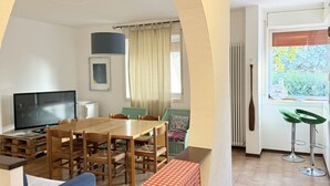 Quarto com mesa de jantar de madeira, cadeiras, televisão, janela com cortinas e radiador.