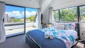 Quarto com uma cama grande, duas janelas e vista para áreas verdes e edifícios.