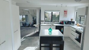 Uma cozinha moderna com armários brancos, uma área de refeições com mesa de vime preto e vista para o exterior por grandes janelas.