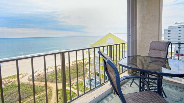 Balcón con mesa y sillas de vidrio con vista a una playa y un edificio.