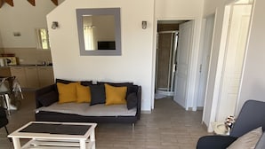 Ein geräumiger Wohnbereich mit einer Couch, einem Couchtisch und einem Sessel. Im Hintergrund ist eine Küche mit Fenster und Spüle zu sehen.