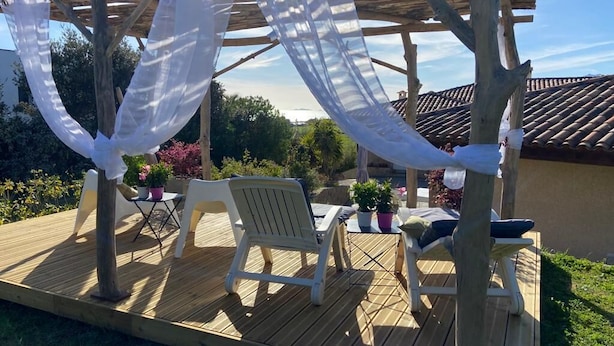 Eine Holzterrasse mit weißen Stühlen und einem Tisch, eine Pergola mit weißem Stoff und ein Ziegeldach im Hintergrund.