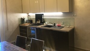 Een kleine keuken met een gasfornuis, een spoelbak en een aanrecht. Boven het aanrecht zijn kastjes en er staat een eettafel met stoelen.