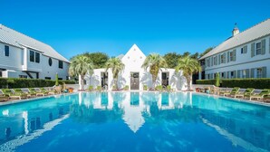 A large swimming pool with lounge chairs and palm trees, surrounded by white buildings with a central structure featuring a clock.