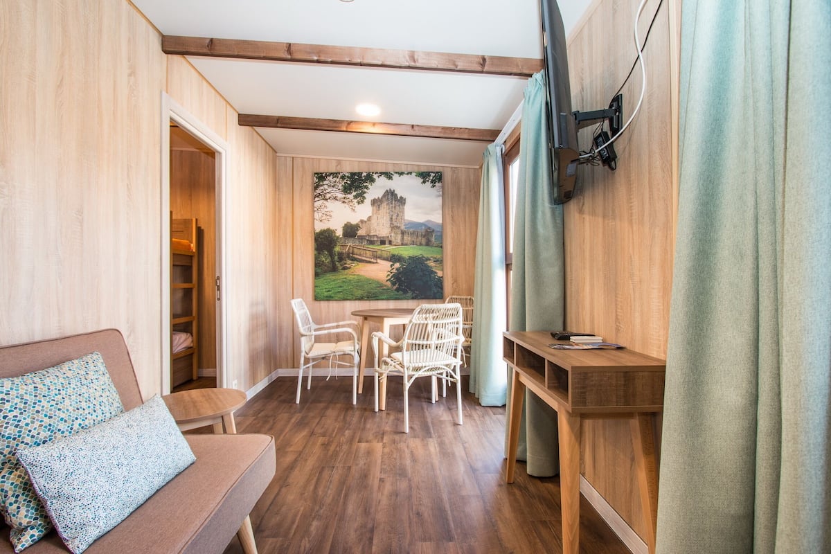 A cozy room with a wooden floor, a dining area with chairs, a table, a sofa with patterned cushions, a painting of a castle, and a TV mounted on the wall.