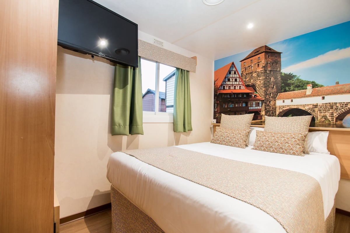 A bedroom with a large bed, green curtains, a flat-screen TV mounted on the wall, and a mural of a medieval building.