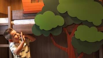 Een stapelbed met een houten hoofdeinde, een klein tafeltje met een afstandsbediening, en een uit papier gesneden afbeelding van een huis en boom.