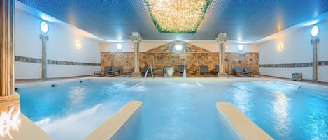 Una amplia piscina interior con dos áreas separadas, un techo con diseño geométrico y una pared de piedra con un arco de piedra.
