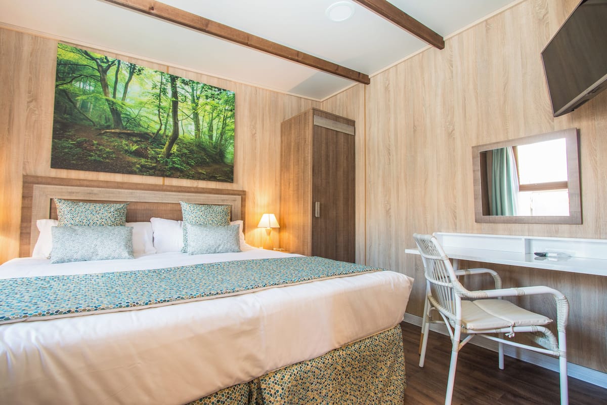 Un dormitorio con cama, escritorio, silla, televisor y un cuadro de un bosque.