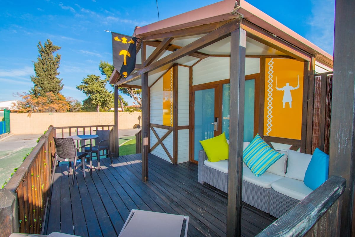 A covered outdoor seating area with a wooden deck, wicker furniture, and a sofa with colorful cushions.