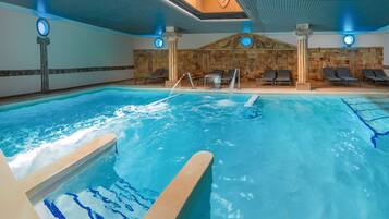 A large indoor swimming pool with a waterfall feature, lounge chairs, and stone walls.