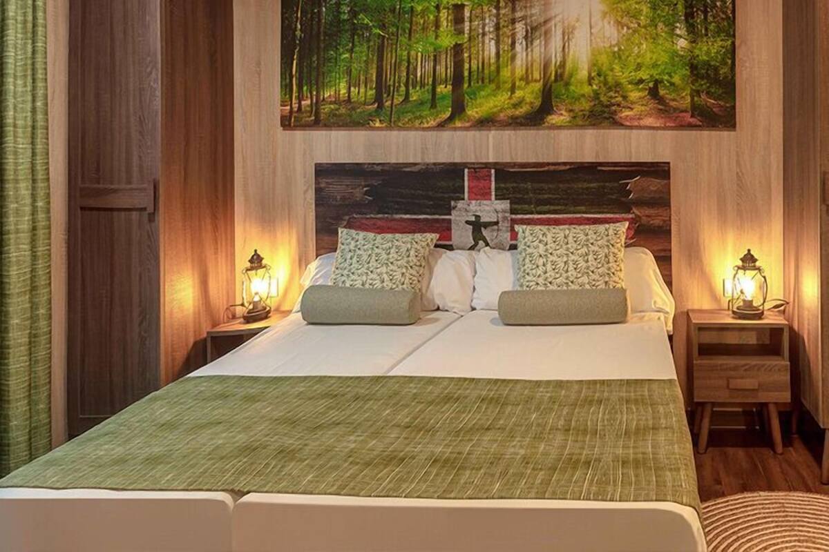 A double bed with a green bedspread, two nightstands with wall-mounted lamps, and a forest-themed painting above the bed.