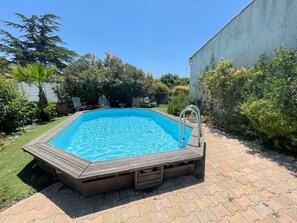 Une piscine rectangulaire dotée d’une terrasse en bois et d’un escalier en métal, entourée d’une végétation luxuriante et d’une terrasse en briques.