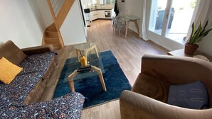 Una sala de estar moderna con sofá, sillón y una mesa de centro de vidrio. Una escalera conduce a una zona de cocina con una mesa de comedor.
