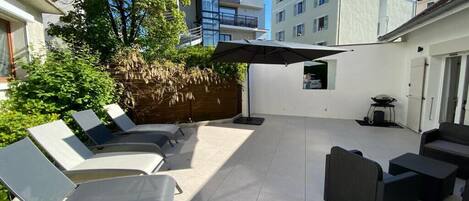 Un espace extérieur aménagé en terrasse, avec des chaises longues et une table à manger, entouré d’un petit muret et d’un grand parasol.