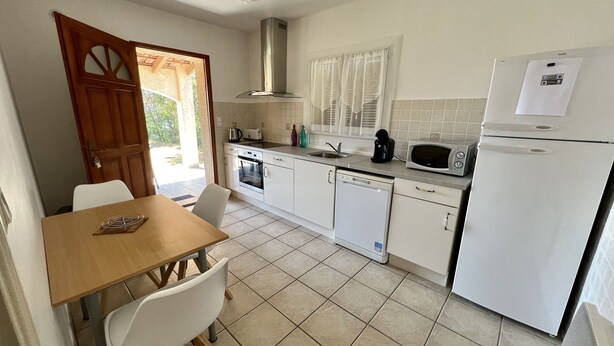 Una cocina compacta con una mesa de madera, sillas blancas, horno empotrado, microondas, lavavajillas y refrigerador.